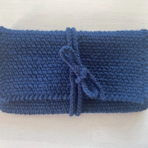 Op de afbeelding: Een marineblauwe gebreide clutch met een flap en een bijpassende striksluiting. De clutch is rechthoekig en heeft een getextureerde uitstraling. De strik is in het midden geknoopt.