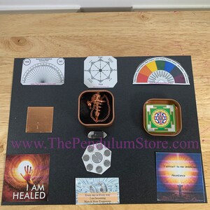 May include: A black background with a variety of pendulum tools and charts. The tools include a pendulum, a chart for numerology and astrology, a color chart, a chart for yes or no questions, and a chart for chakras. The charts are printed on white paper and have black text. The background is black and has the text "www.ThePendulumStore.com" printed in white.