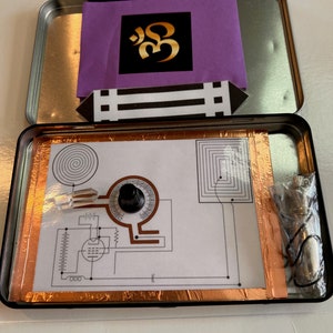 May include: A silver metal box with a purple paper containing a gold Om symbol. The box is open and reveals a circuit board with copper tape, a knob, and a crystal.