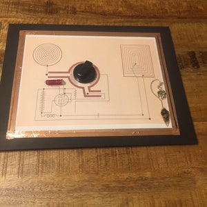May include: A framed copper-toned pendulum board with a black knob, red coils, and a black and gold pendulum hanging from the top. The board features a spiral, a square, and a circuit diagram. The text "www.ThePendulumStore.com" is printed at the bottom.