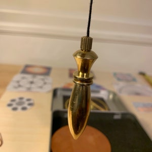 Peut inclure: Un pendule en laiton avec une pointe pointue et une ficelle noire suspendue à un plafond. Le pendule est suspendu au-dessus d'une surface circulaire brune.