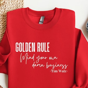 Könnte beinhalten: Ein leuchtend rotes Sweatshirt mit dem weißen Text "GOLDEN RULE Mind your own damn business -Tim Walz-". Das Sweatshirt ist ordentlich gefaltet und präsentiert die Botschaft. Das Design ist schlicht und die Botschaft ist deutlich.