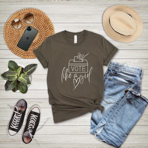 May include: An olive green t-shirt with the text "Vote like a girl" in white script, featuring a ballot box graphic. The shirt is styled with blue jeans, a straw hat, sunglasses, a smartphone, a small potted plant, and black and white sneakers.