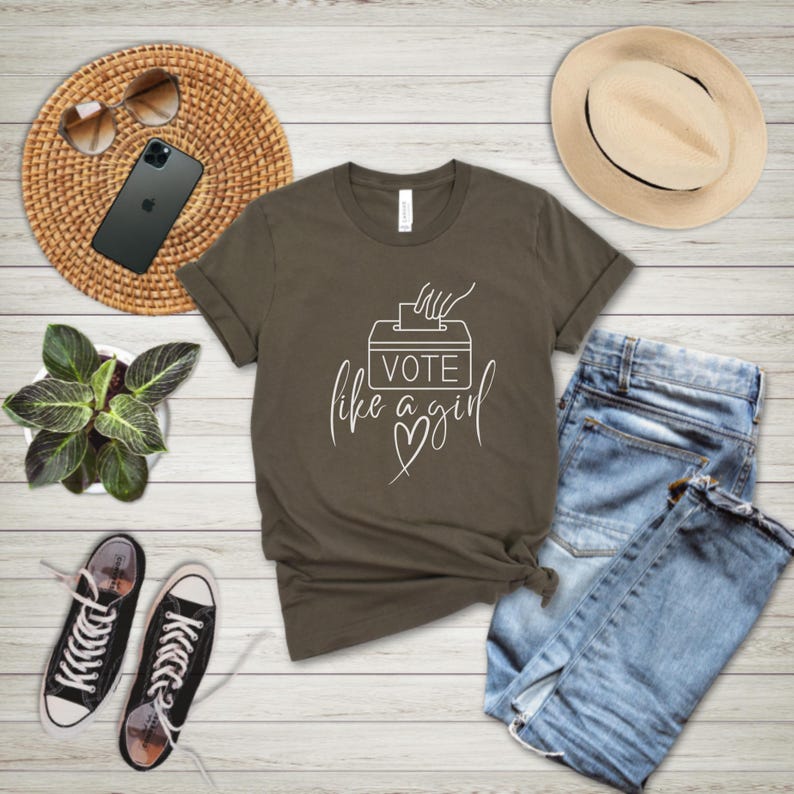 May include: An olive green t-shirt with the text "Vote like a girl" in white script, featuring a ballot box graphic. The shirt is styled with blue jeans, a straw hat, sunglasses, a smartphone, a small potted plant, and black and white sneakers.