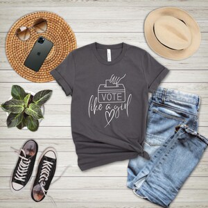 May include: A charcoal gray t-shirt with the text "VOTE like a girl" in white script, paired with blue jeans, black sneakers, and a straw hat. The image also includes a smartphone, sunglasses, and a small potted plant.