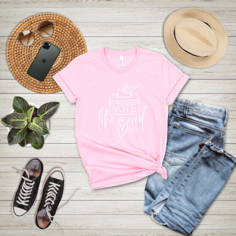 May include: A light pink t-shirt with the text "VOTE like a girl" in white script. The shirt is paired with blue jeans, black sneakers, a straw hat, sunglasses, and a smartphone on a woven mat.