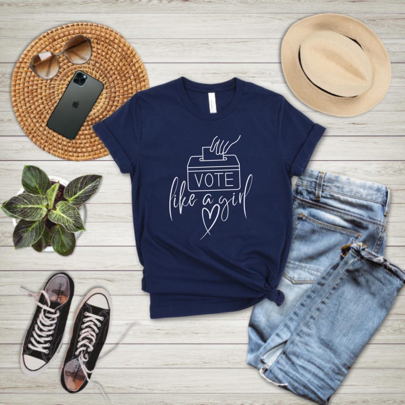 May include: Navy blue t-shirt with the text "Vote like a girl" in white script. The shirt is paired with blue jeans, black and white sneakers, a straw hat, and sunglasses. A smartphone and a small potted plant are also visible.