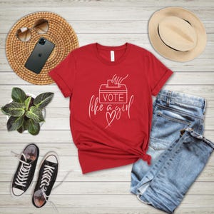 May include: A red t-shirt with the text "Vote like a girl" in white script. The design includes a ballot box and a heart. The shirt is paired with blue jeans, black sneakers, and a straw hat.