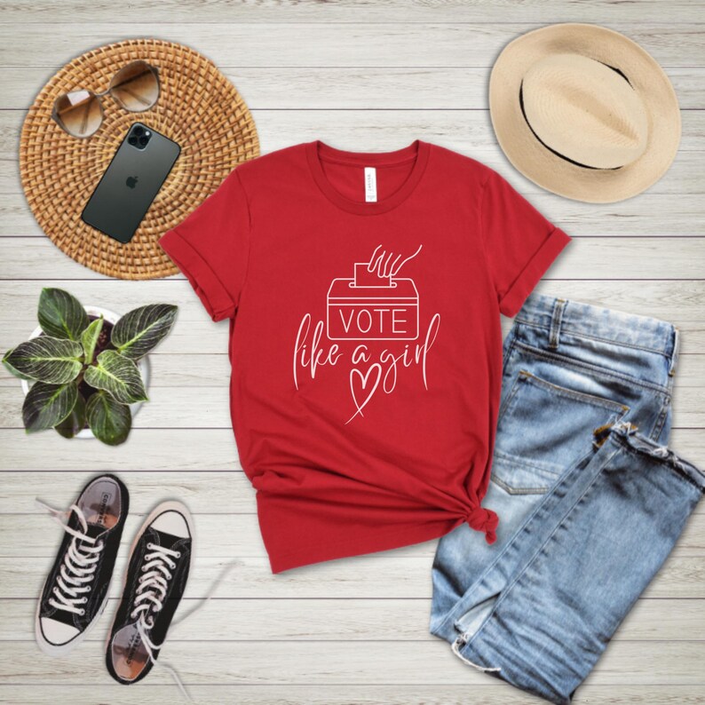 May include: A red t-shirt with the text "Vote like a girl" in white script. The design includes a ballot box and a heart. The shirt is paired with blue jeans, black sneakers, and a straw hat.