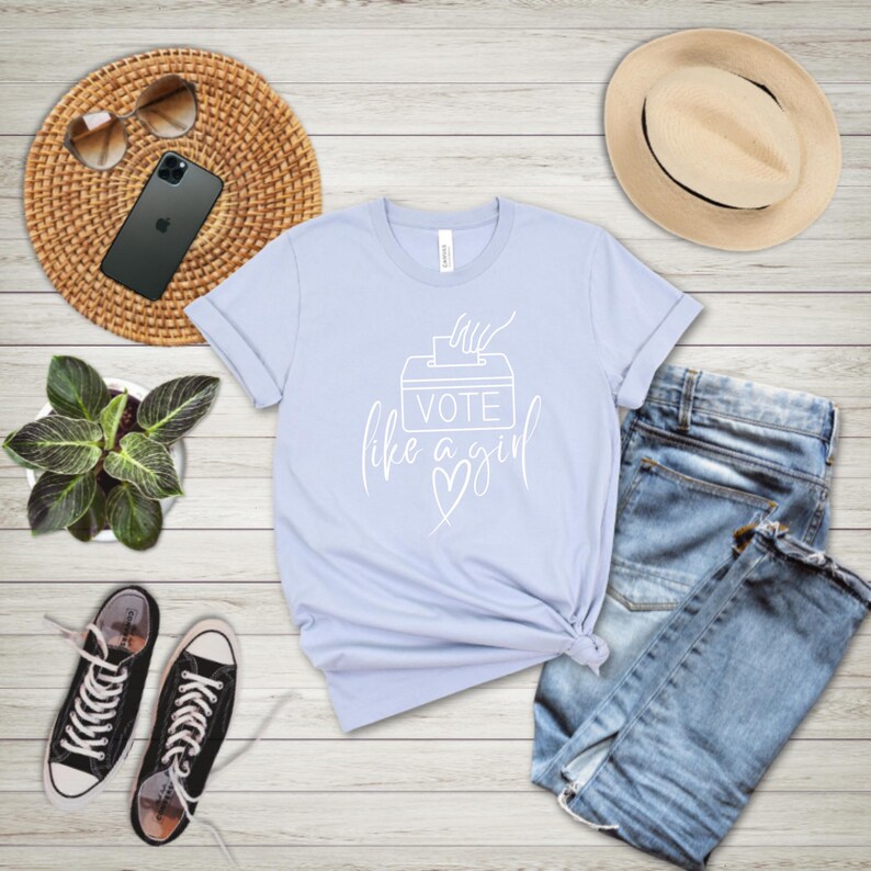 May include: A light blue t-shirt with the text "VOTE like a girl" in white script. The shirt is styled with blue jeans, black sneakers, a straw hat, sunglasses, a smartphone, and a small potted plant.