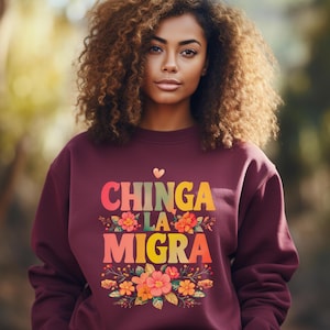 May include: Burgundy sweatshirt with the text "CHINGA LA MIGRA" in colorful letters, a heart, and floral accents. The design is centered on the front of the sweatshirt.
