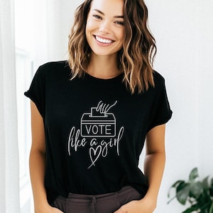 May include: Black t-shirt with the text "VOTE like a girl" in white script. The design includes a ballot box and a heart. The shirt is worn by a person with brown hair.