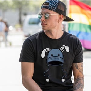 Può includere: T-shirt nero con un design a cartoni animati di un berretto blu con orecchie da coniglio bianche e pompon. La persona indossa un cappellino da camionista a scacchi neri e blu e occhiali da sole.