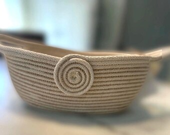 May include: A beige woven rope basket with a circular woven rope handle. The basket is oval shaped and has a wide opening.