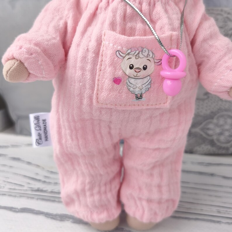 May include: Handmade doll in pink jumpsuit with pocket and sticker on it. There is a tag ,, Cutedollshandmade,,on the romper. The doll has a silver necklace with a mini pacifier. The doll is approximately 20 cm tall.