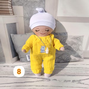8. May include: A handmade doll wearing a bright yellow jumpsuit with a hippo patch and a white cap.  The doll has closed eyes and a smiling expression. A silver necklace with a white pendant is around the doll's neck. The doll is approximately 8 inches tall.