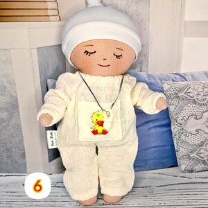 6. May include: Handmade doll in milky overalls with a yellow chick sticker and a white hat. The doll has closed eyes and a smiling face. The doll wears a silver necklace with a white pendant. The doll is approximately 20 cm tall.
