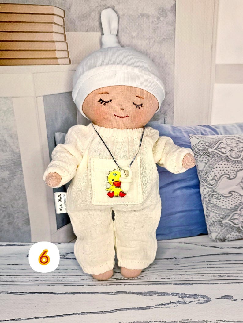 6. May include: Handmade doll in milky overalls with a yellow chick sticker and a white hat. The doll has closed eyes and a smiling face. The doll wears a silver necklace with a white pendant. The doll is approximately 20 cm tall.