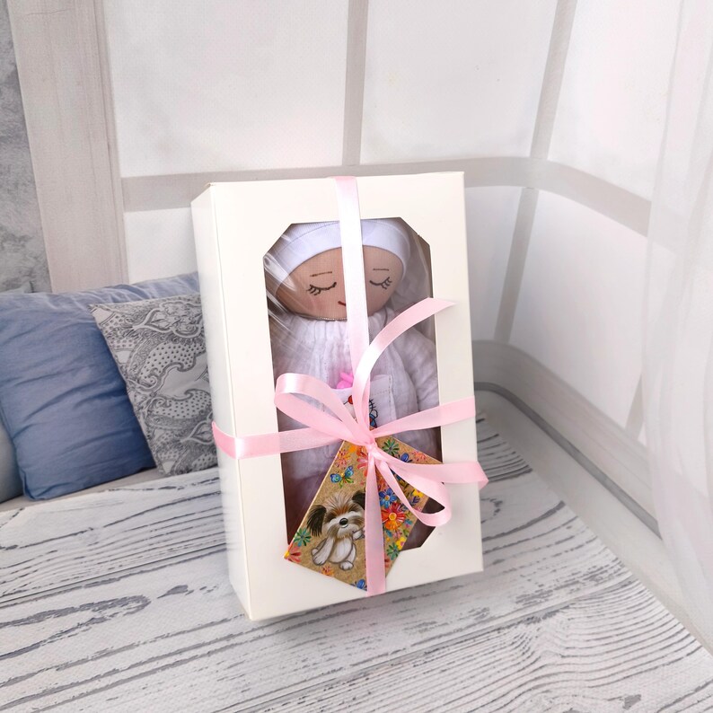 May include: A handmade doll in a white box with a clear window, tied with a pink ribbon. The doll is wearing a white bonnet and dress. A small tag with a dog illustration is attached to the ribbon. The background includes pillows and a window.