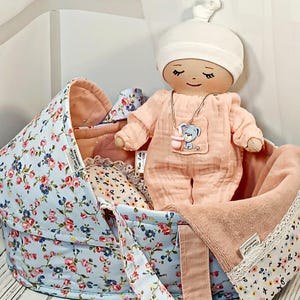 Puede incluir: Una muñeca hecha a mano con un mono color melocotón y un gorro blanco descansa en un portabebés con estampado floral. El portabebés es azul claro con un diseño floral rosa. Dentro hay una manta y una almohada a juego.