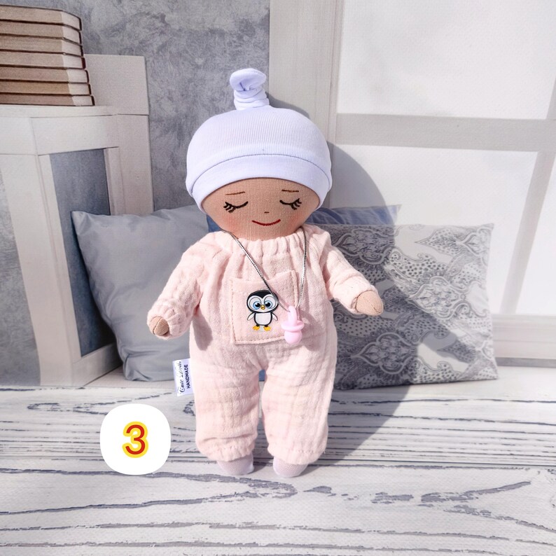 3. May include: A handmade doll wearing a light pink jumpsuit and a white cap. The doll has a small pocket with a penguin design and a pink pacifier. The doll is standing on a wooden surface.