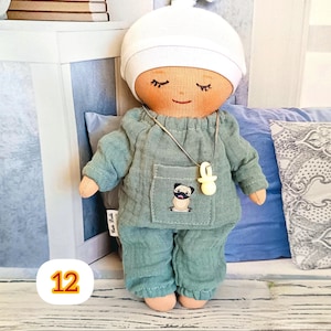 12. May include: A handmade doll wearing a light teal pajama set and a white cap. The doll has closed eyes, a smiling mouth, and a necklace with a pacifier pendant. A pug is embroidered on the pocket.