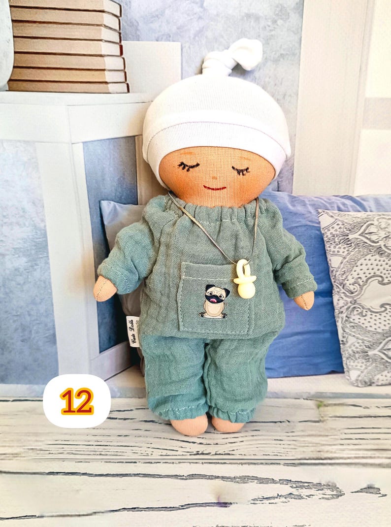12. May include: A handmade doll wearing a light teal pajama set and a white cap. The doll has closed eyes, a smiling mouth, and a necklace with a pacifier pendant. A pug is embroidered on the pocket.