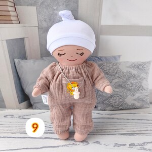 9. May include: Handmade doll wearing beige jumpsuit with lion patch and white hat. Doll has closed eyes and smiling face. Doll wears silver necklace with white pendant. Doll is approximately 20 cm tall.