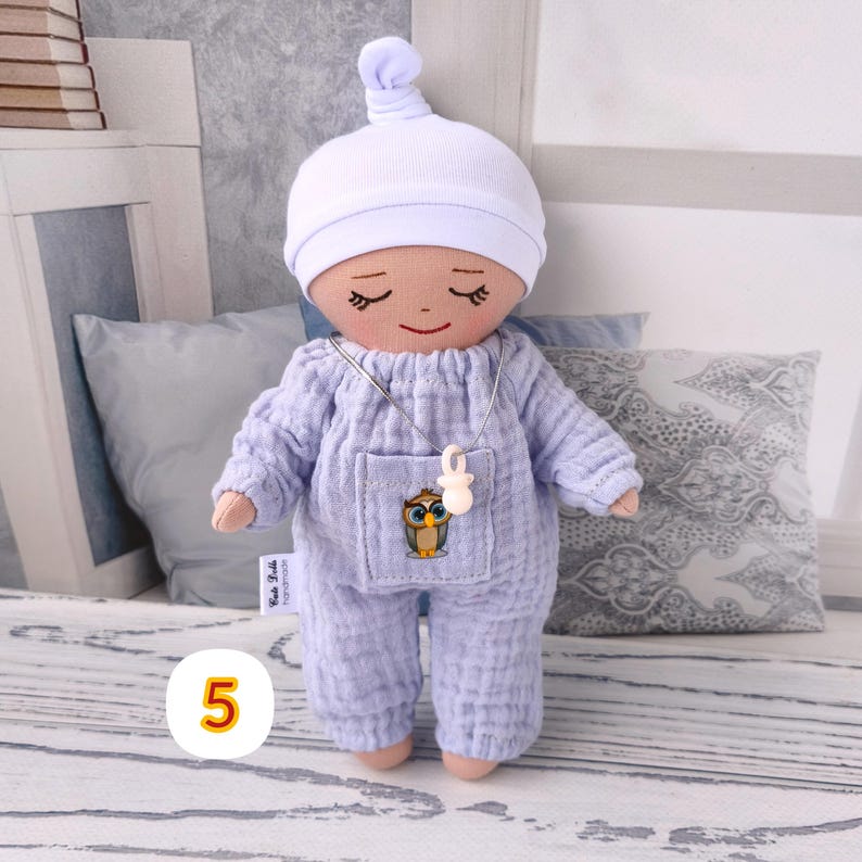 5. May include: Handmade doll wearing lavender jumpsuit with owl sticker and white hat. Doll has closed eyes and smiling face. Doll wears silver necklace with white pendant. Doll measures approximately 20 cm tall.