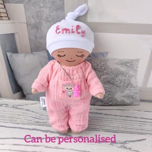 May include: A handmade doll wearing a pink onesie and a white cap embroidered with the name "Emily" in pink. The doll has a necklace with a pink pacifier pendant and a pocket with a sheep graphic. The text "Can be personalised" is at the bottom.