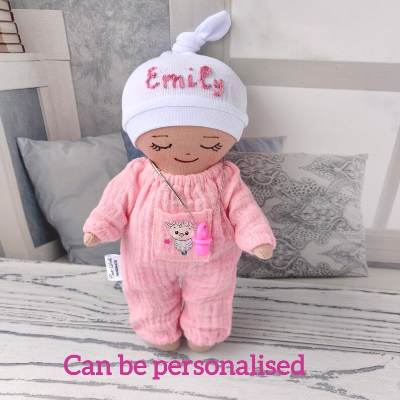 May include: A handmade doll wearing a pink onesie and a white cap embroidered with the name "Emily" in pink. The doll has a necklace with a pink pacifier pendant and a pocket with a sheep graphic. The text "Can be personalised" is at the bottom.