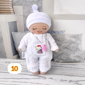 10. May include: A handmade doll wearing a white onesie, matching cap, and a pink pacifier necklace. The doll has a smiling face with closed eyes and a small pocket with a Hello Kitty graphic. The doll is approximately 8 inches tall.