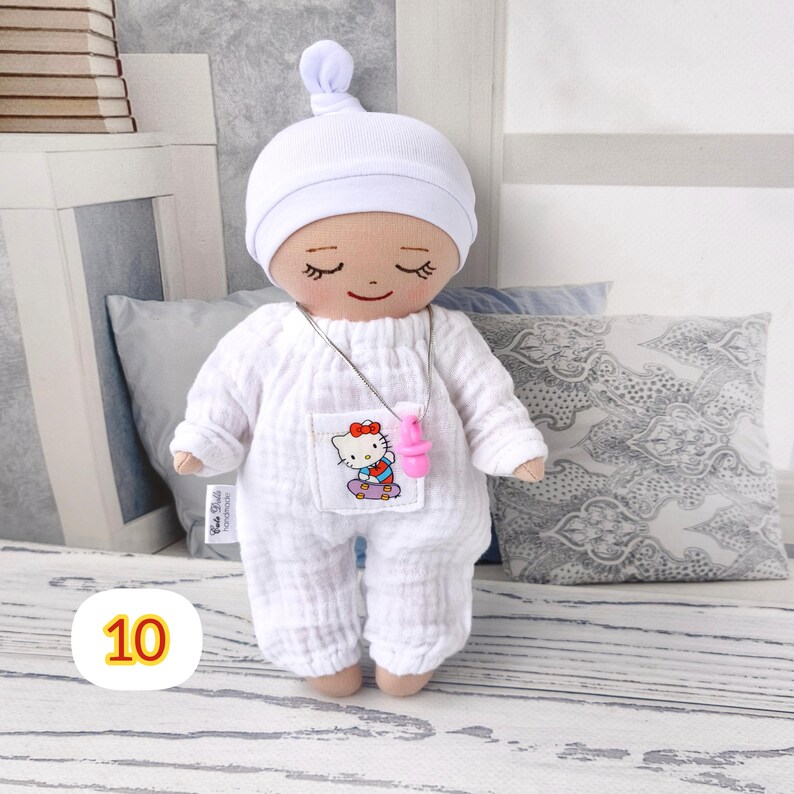 10. May include: A handmade doll wearing a white onesie, matching cap, and a pink pacifier necklace. The doll has a smiling face with closed eyes and a small pocket with a Hello Kitty graphic. The doll is approximately 8 inches tall.