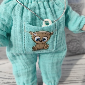 The baby doll is wearing a muslin romper with a pocket. There is a small animal sticker on the pocket. A mini pacifier is placed in the pocket.