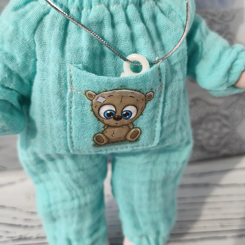The baby doll is wearing a muslin romper with a pocket. There is a small animal sticker on the pocket. A mini pacifier is placed in the pocket.