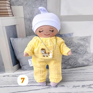 7. May include: Handmade doll wearing a yellow jumpsuit with a horse sticker and a white hat. The doll has closed eyes and a smiling face. The doll wears a silver necklace with a white pendant. The doll is approximately 20 cm tall.