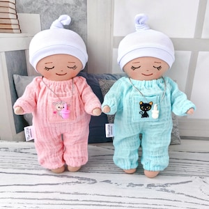 Two baby dolls with mini pacifiers to choose from. One doll is dressed in a pink romper with a pocket and a white hat, the other is dressed in a pale blue jumpsuit and a white hat. The jumpsuit has a small pocket with a sticker.