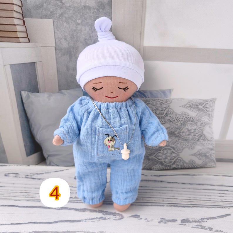 4. May include: A handmade doll wearing a light blue onesie and a white cap. The doll has closed eyes and a small embroidered dog on the pocket. A silver necklace with a pacifier pendant hangs around its neck. The doll is a soft toy.