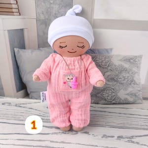 1. May include: Handmade doll wearing pink jumpsuit with sheep patch and white hat. Doll has closed eyes and smiling face. Doll wears silver necklace with white pendant. Doll is approximately 20 cm tall.