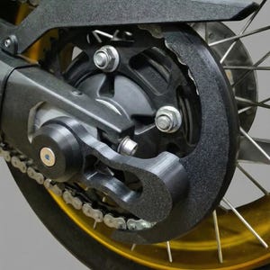 Op de afbeelding: Detailopname van de achterwielconstructie van een motorfiets. De afbeelding toont de zwarte kettingbeschermer, het tandwiel en de ketting tegen een goudkleurige velg. De band is zwart en de spaken zijn zilver.