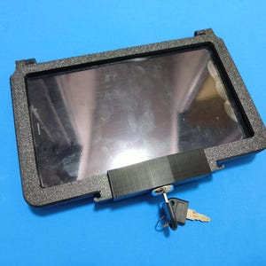 Puede incluir: Una carcasa negra para tableta con mecanismo de cierre y dos llaves. La carcasa tiene una superficie texturizada y una abertura rectangular para la pantalla de la tableta. Las llaves están unidas a un pequeño llavero.