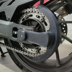 May include: Close-up of a motorcycle's rear wheel assembly. The image highlights the chain, sprocket, and a black chain guard. The tire has the brand name "Michelin" visible. The assembly is primarily gray and black.