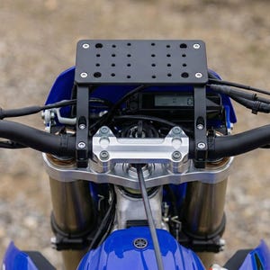 May include: Close-up of a motorcycle handlebar assembly. The image shows a black mounting bracket with multiple holes, attached to the handlebars. The motorcycle has a blue fairing and a silver handlebar clamp.