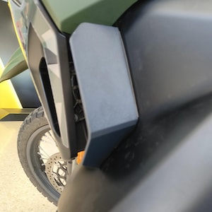 Op de afbeelding: Een close-up van een motorfiets carrosseriepaneel met een mat olijfgroen bovengedeelte en een mat zwart ondergedeelte. Een donkergrijze hoekige plastic luchtinlaatkap is aan de zijkant gemonteerd, gepositioneerd nabij het voorwiel.