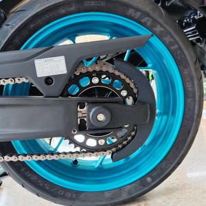 Peut inclure: Une roue arrière de moto dotée d'une jante cyan vif et d'un pneu noir avec la marque Maxxis visible sur le flanc. L'ensemble comprend un protège-chaîne en métal noir, une chaîne de transmission métallique et une couronne noire avec le nombre 412.
