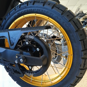 Op de afbeelding: Close-up van een motorwiel met een zwarte band en een gouden velg. De band heeft de tekst "SCORPION RALLY" erop gedrukt. Het wiel is bevestigd aan een zwart frame met een ketting en tandwiel.