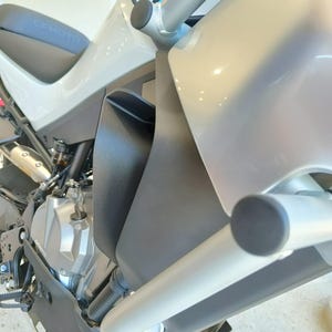 May include: A side view of a CFMOTO motorcycle featuring a light gray fuel tank, a black seat with the CFMOTO logo, a matte black side fairing, and silver tubular crash bars. The engine block is visible with metallic components and black piping.