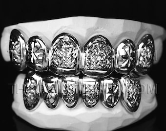 May include: Silver-colored grillz set on a white dental mold. The grillz have a geometric, diamond-cut design, reflecting light. The image is set against a black background, highlighting the metallic shine of the teeth.