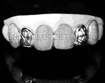 May include: Close-up of a set of silver grillz with a diamond-like texture. The grillz are designed to fit over the front teeth and have a decorative pattern.