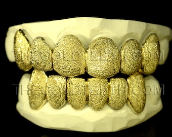 Może przedstawiać: Złote grillzy na białym odlewie dentystycznym. Grillzy mają teksturowaną powierzchnię i są prezentowane na czarnym tle. Tekst "THEGOLDTEETH.COM" jest widoczny na grillzach.